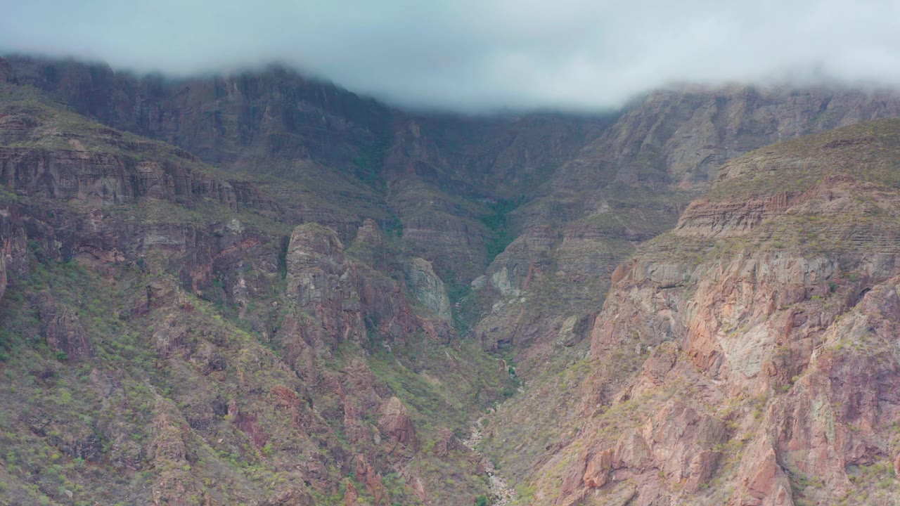 A high view of the Puerto Escondido mountain valley shows a central rocky ridge, darker canyon walls behind it, low cloud on the peaks, and no visible people.