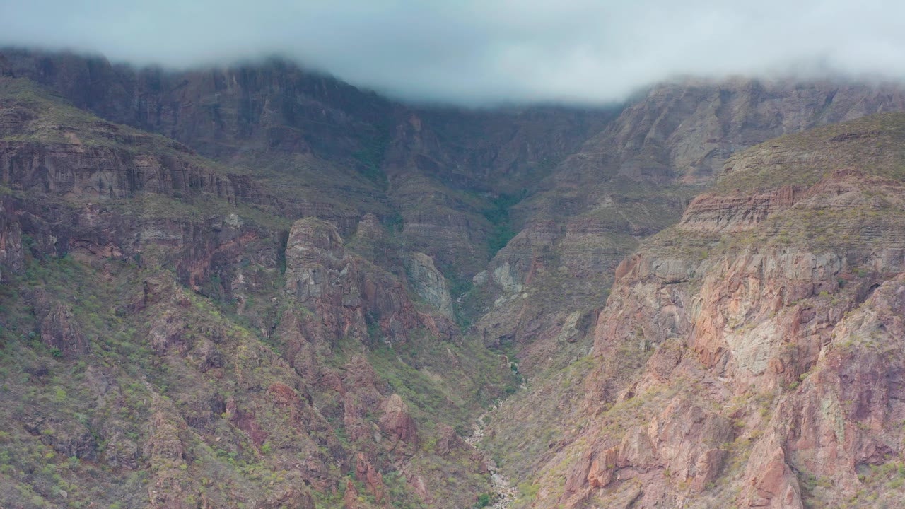 Layered red-brown and gray slopes in Puerto Escondido sit below misty mountain tops, with small green patches across the terrain and no visible people.