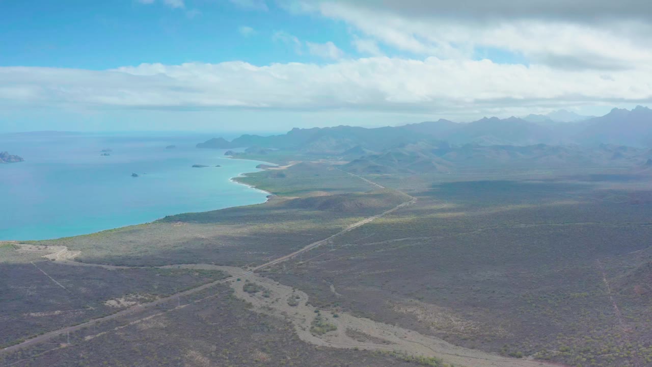 Aerial frame over Puerto Escondido with dry brown terrain in the foreground, intersecting dirt tracks, turquoise coastal water, hazy mountain ridges, cloud bands, and no visible people.