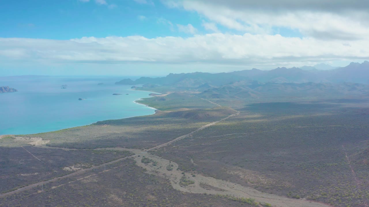 Puerto Escondido aerial frame with broad desert flats in the foreground, a curved sandy coastline at left, blue ocean beyond, layered mountains on the horizon, and no visible people.