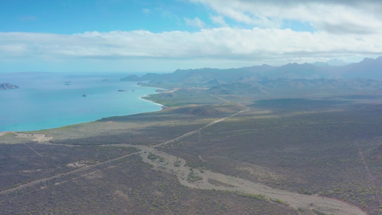 Aerial view of dirt road, arid terrain, turquoise ocean, and distant mountains.