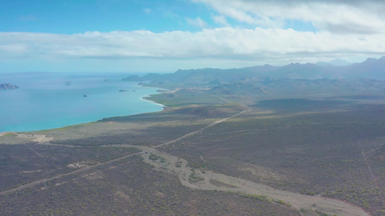 A slightly later aerial view of Puerto Escondido shows the pale road running diagonally through scrubland toward the coastline, with turquoise water and mountain ridges beyond, and no visible people.
