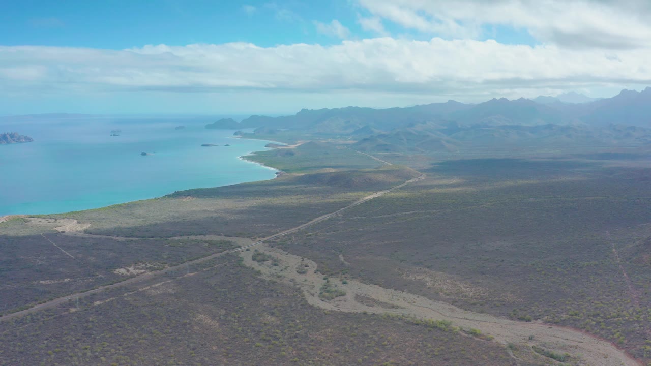 The aerial view continues over Puerto Escondido with more of the coastline visible at left, a dirt road crossing the brown scrubland, hazy mountain silhouettes in the distance, and no visible people.