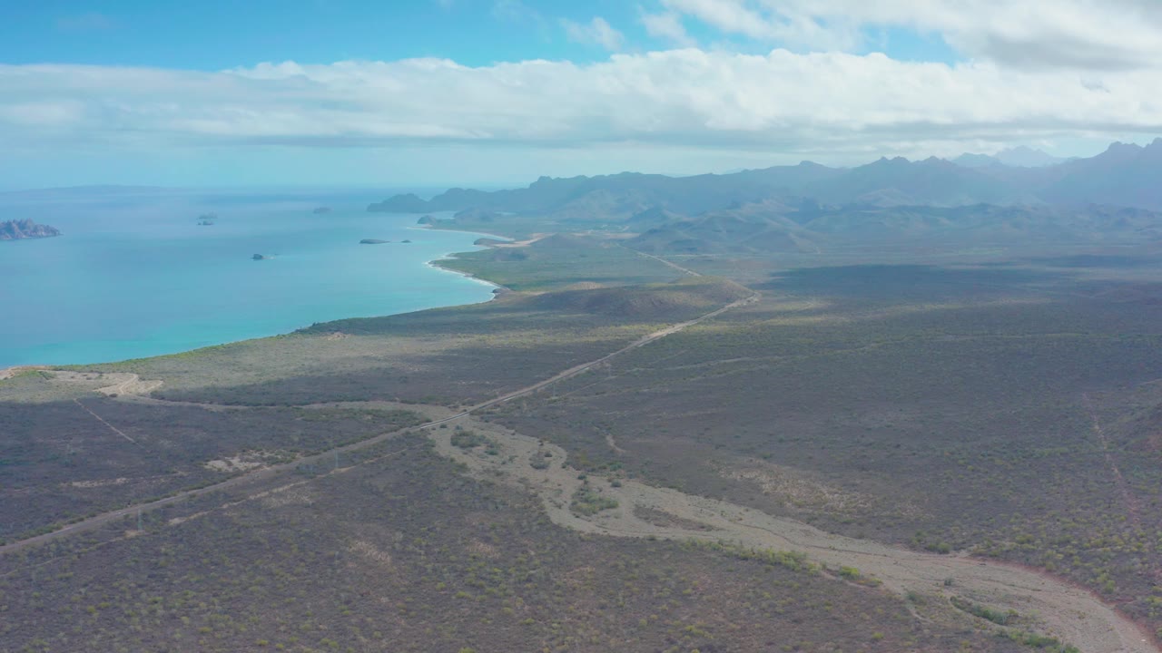 Mid-clip, the frame shows Puerto Escondido's turquoise bay along the left edge, scrubby flat terrain and a winding dry wash below, a dirt road through the middle distance, and no visible people.