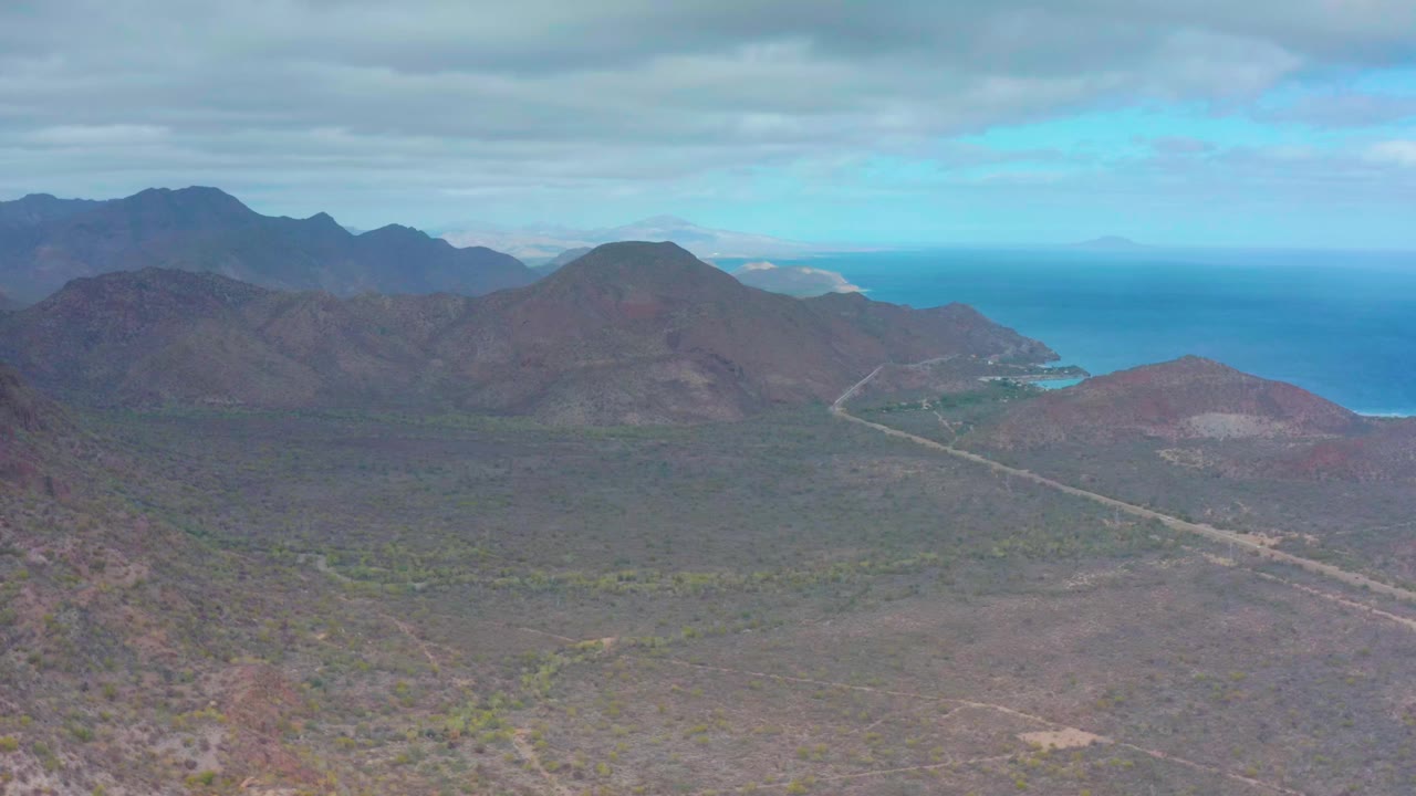 At 0:00, an elevated view over Puerto Escondido, Baja California Sur shows rocky brown mountains, sparse vegetation, a pale road toward the coast, blue ocean, gray clouds, and no visible people.