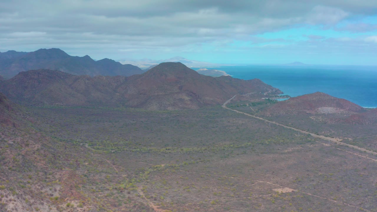 Opening frame over Puerto Escondido, Baja California Sur, showing dry mountain slopes, a valley road, distant blue ocean, overcast sky, and no visible people.