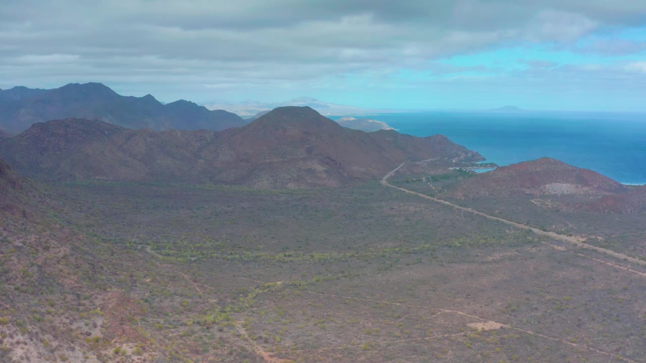Early frame over Puerto Escondido, Baja California Sur, with the pale road more centered through the dry valley, mountain ridges on both sides, ocean beyond, and no visible people.