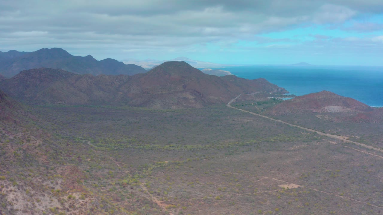 Wide aerial view over Puerto Escondido, Baja California Sur, with rocky desert slopes, a broad shrub-covered valley, distant mountains, blue ocean, cloud cover, and no visible people.
