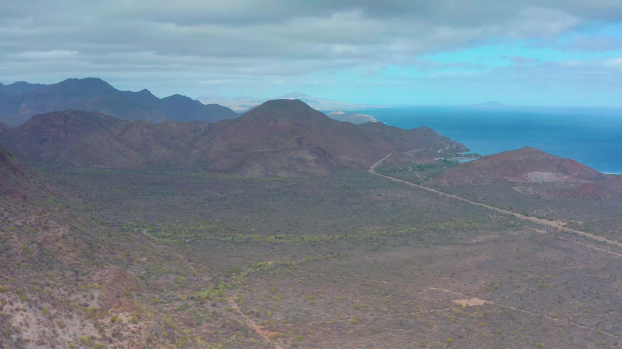 Aerial frame over Puerto Escondido, Baja California Sur, showing a pale road through the arid valley floor, dark mountain ridges, the ocean beyond the coast, overcast sky, and no visible people.