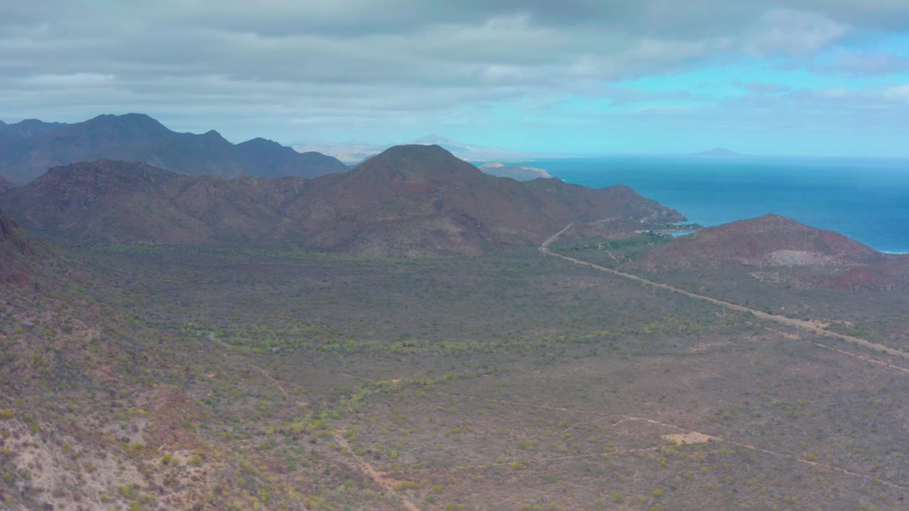 Dry valley and dark mountain ridge beneath a cloudy sky with ocean in the distance.