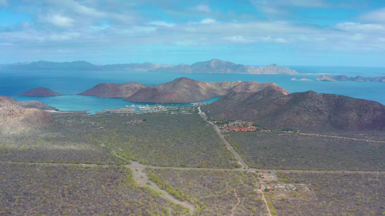 Aerial frame at 0:01 over Puerto Escondido showing intersecting roads across dry green-brown scrub, a small settlement near the water, rounded hills, blue bay, and no visible people.
