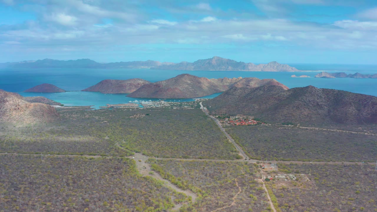 Aerial frame from Puerto Escondido, Baja California Sur, with the dirt road leading through scrubland toward low mountains, turquoise coastal water, and no visible people.