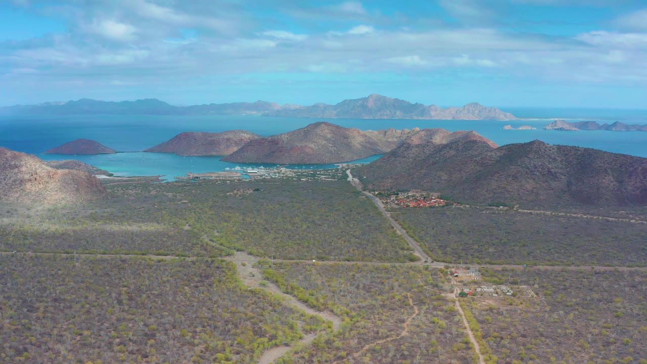 Aerial frame in Puerto Escondido BCS showing pale dirt tracks through dry scrub, rounded coastal hills, turquoise shoreline water, distant landforms, and no visible people.