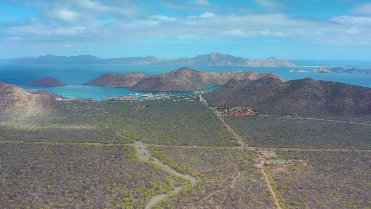 Puerto Escondido BCS aerial frame with a diagonal dirt road cutting through arid scrub, coastal hills across the center, bright blue bay water behind them, and no visible people.