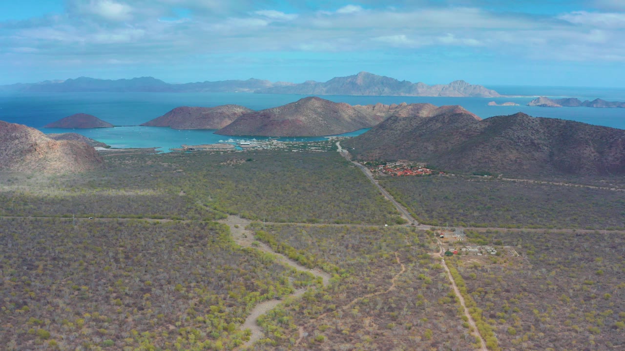 Aerial view over Puerto Escondido, Baja California Sur with scrub desert in front, winding pale roads, rocky hills, blue bay water, distant landforms, and no visible people.
