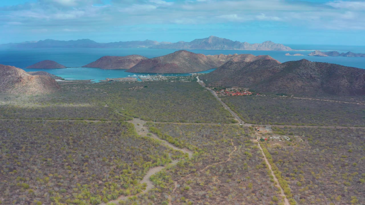 Drone-style view across scrubland toward rocky hills and a blue bay.