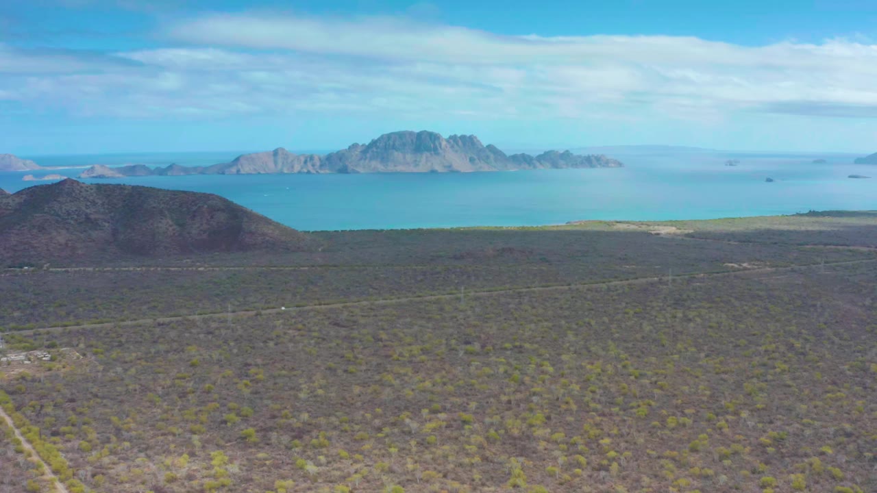 Aerial view over sparse desert vegetation near Puerto Escondido, Baja California Sur, with blue ocean and distant rocky mountains under light cloud, no visible people.