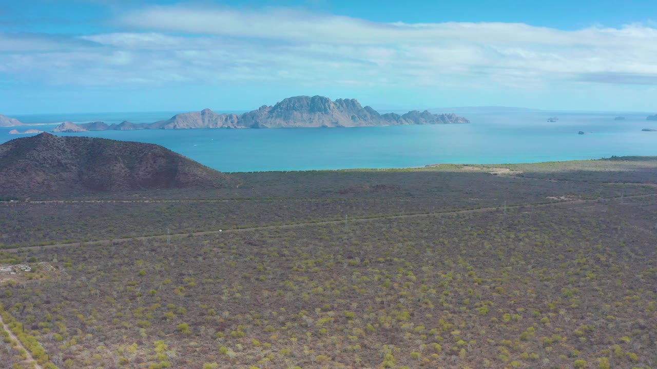 Aerial desert view near Puerto Escondido, Baja California Sur, with scrub vegetation in front, calm blue water across the middle distance, and rocky island-like mountains beyond, no visible people.