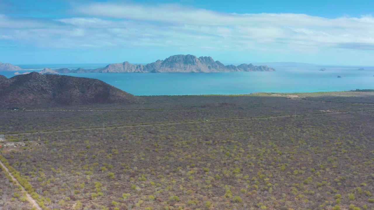 High aerial view across scrub plain toward blue ocean and distant mountains.