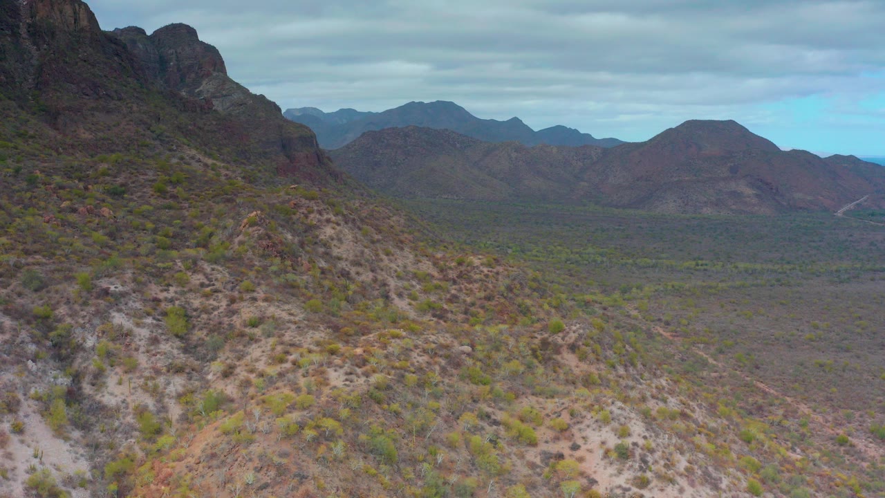 Aerial view over rocky desert slopes and sparse shrubs in Puerto Escondido, Baja California Sur, with layered mountains in the distance and no visible people.