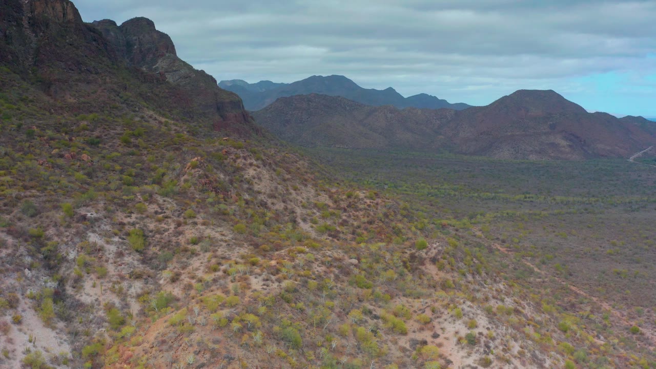 The Puerto Escondido aerial view shows a rocky shrub-covered foreground descending toward a flat desert valley, with layered mountains under gray-blue clouds and no visible people.