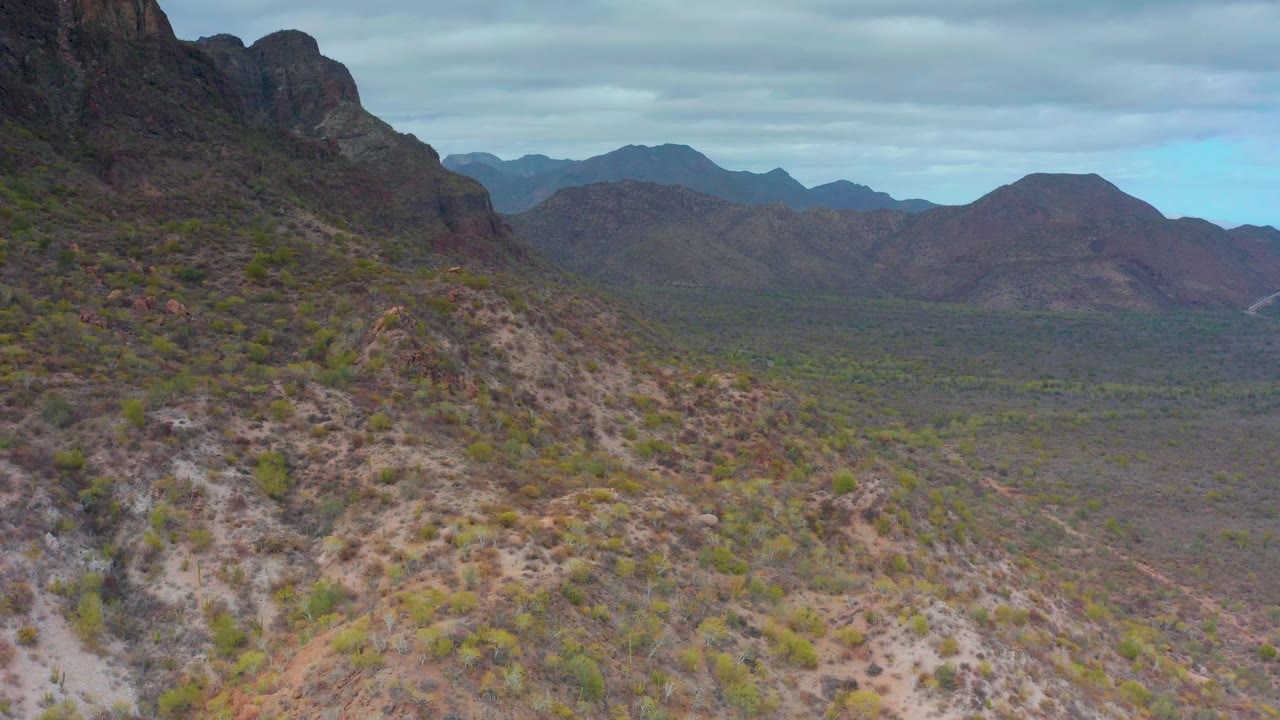 Shrub-dotted hillside and distant brown mountains beneath a cloudy sky.
