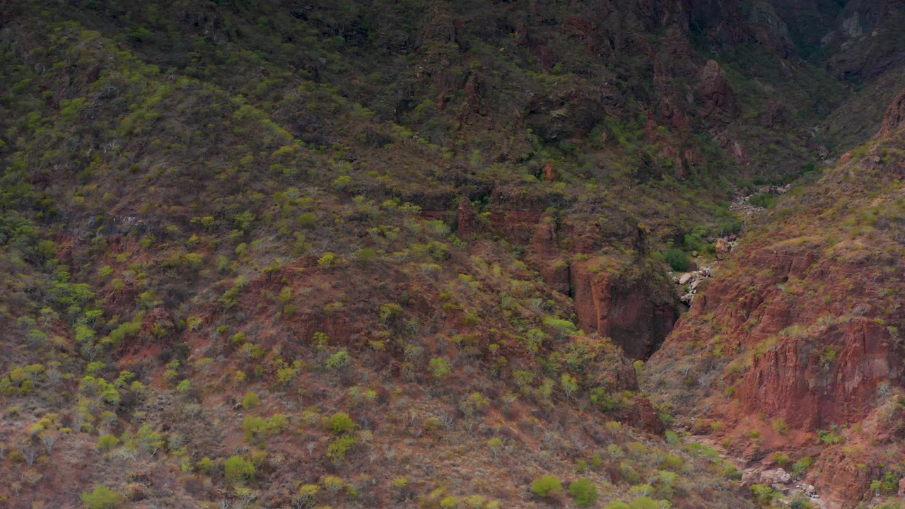 Mid-clip view of the Puerto Escondido canyon, showing steep red and brown slopes, scattered green vegetation, a narrow valley floor, and no visible people.