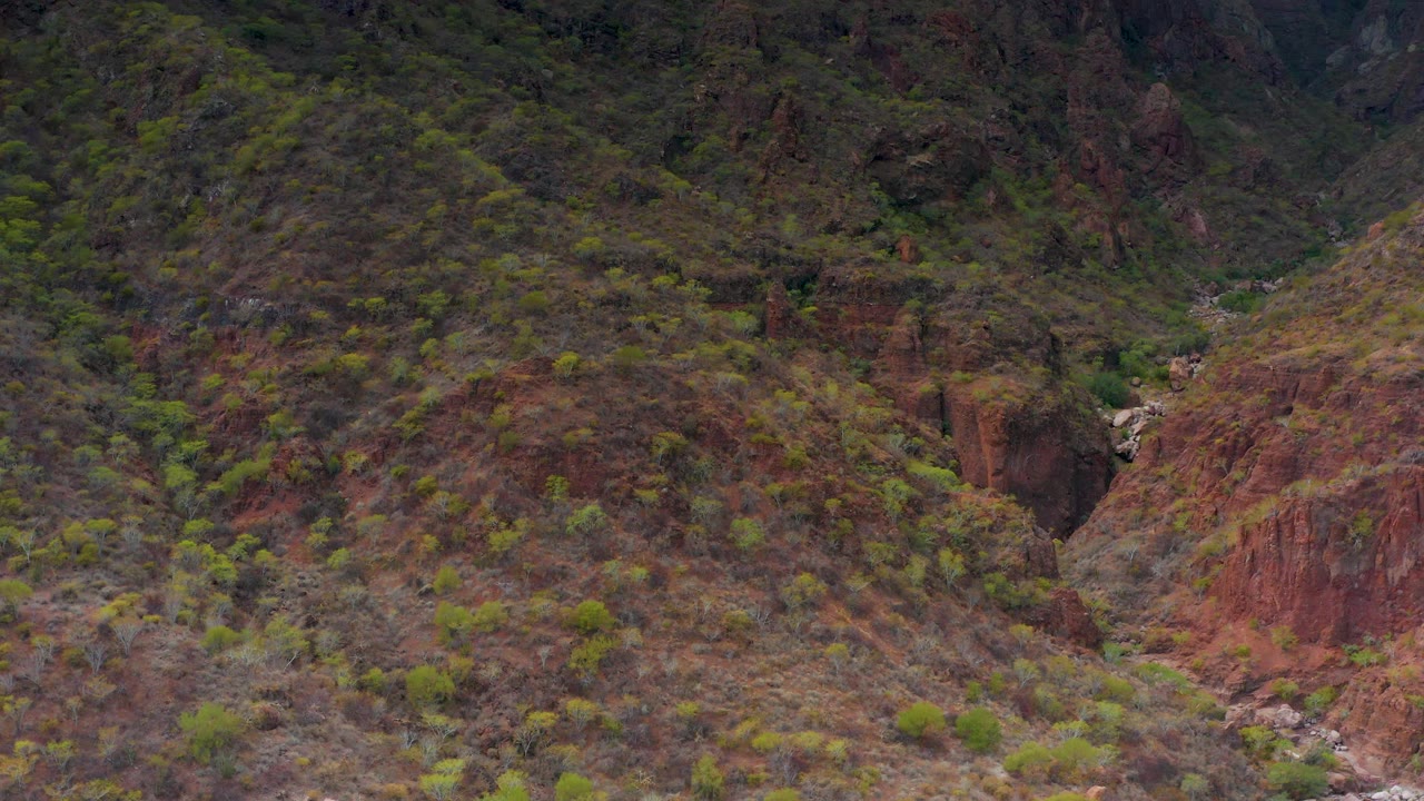 Puerto Escondido mountain terrain shows a reddish-brown slope, scattered green vegetation, a shadowed ravine, and a pale rocky river channel with no visible people.