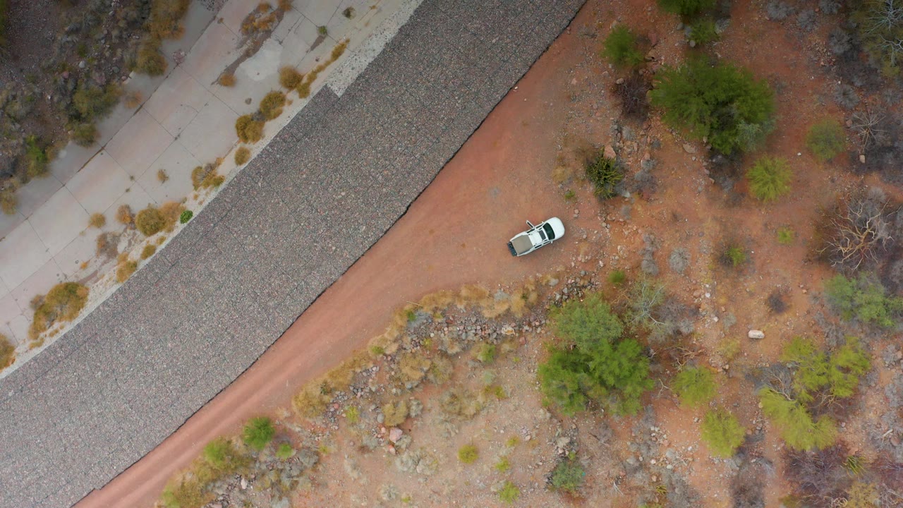Overhead view in Puerto Escondido, Baja California Sur with the white car slightly lower along the red dirt road next to the paved highway, dry brush, rocks, and no visible people.