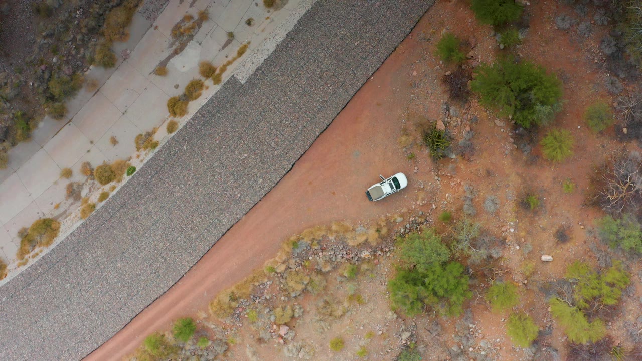 Aerial frame from Puerto Escondido, Baja California Sur showing the white car farther down the red dirt road, with gray highway pavement, pale sidewalk-like roadside areas, shrubs, and no visible people.