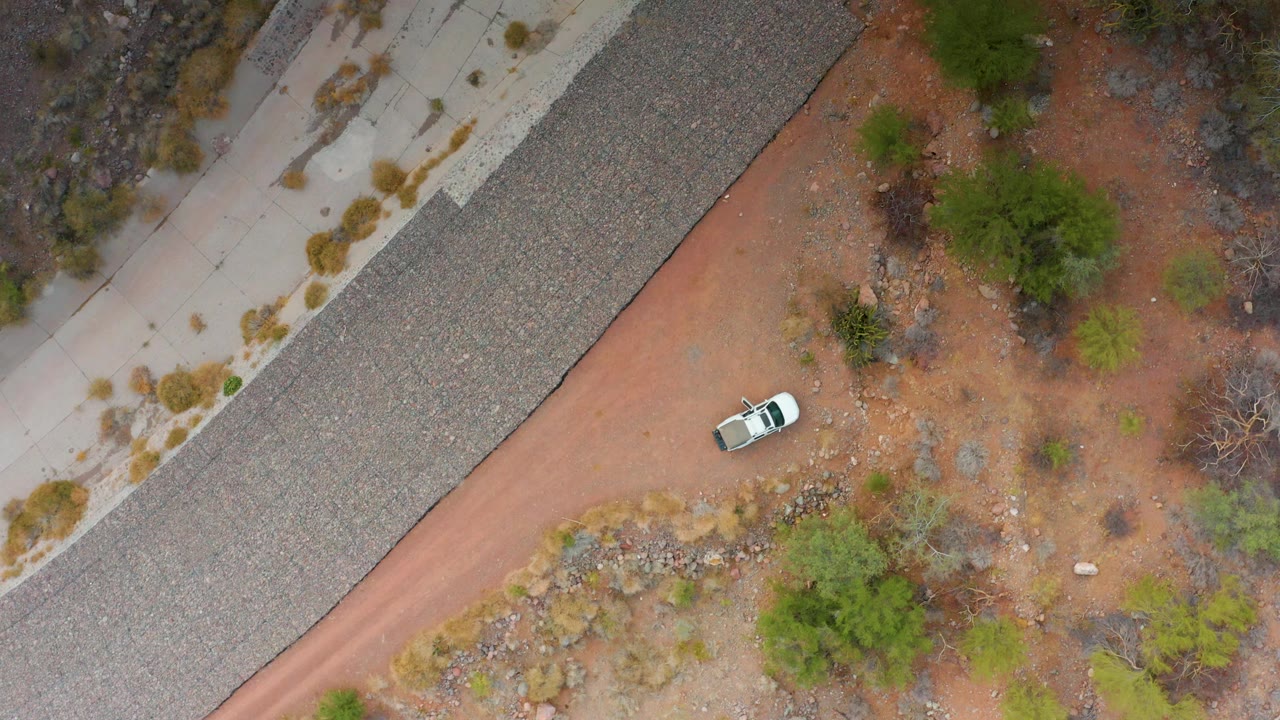Overhead Puerto Escondido, Baja California Sur frame with the white car near the middle of the red dirt road, bordered by a paved highway, scrubby trees, rocky ground, and no visible people.