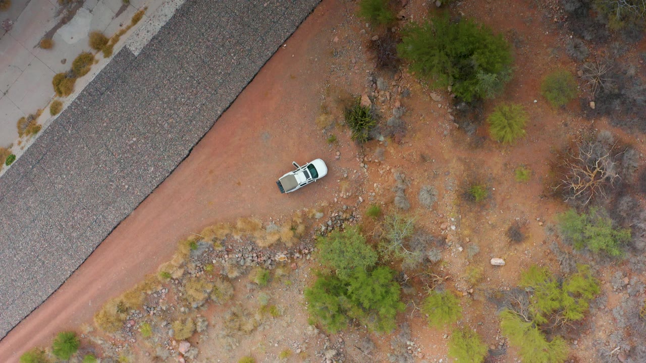 A white car sits farther along the red dirt road in Puerto Escondido, Baja California Sur, framed by rocky ground, scattered bushes, gray gravel, and no visible people.