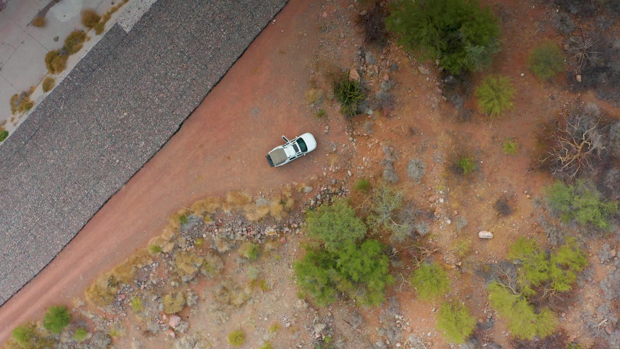 The white car moves near the upper road edge in Puerto Escondido, Baja California Sur, with red dirt, gray gravel, sparse green shrubs, dry brush, and no visible people.