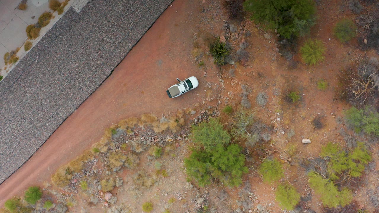 A steep aerial frame shows the white car on a curving red dirt road in Puerto Escondido, Baja California Sur, beside gravel, scrubby vegetation, rocks, and no visible people.
