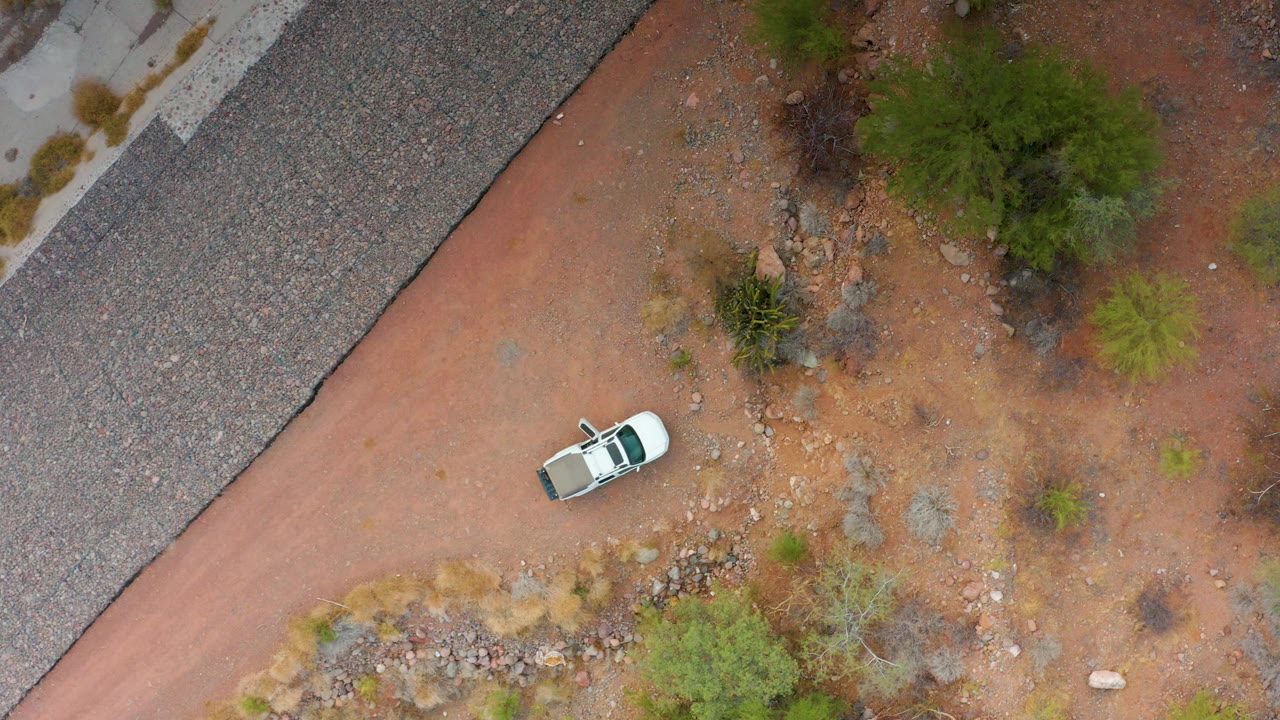 White car on a reddish dirt path beside a paved road in dry rocky terrain.
