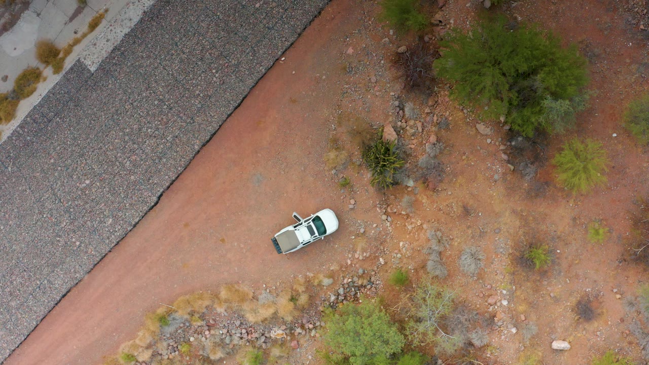 Aerial frame from Puerto Escondido, Baja California Sur, showing the white car slightly farther along the reddish dirt path, with the paved road at the upper left and no visible people.