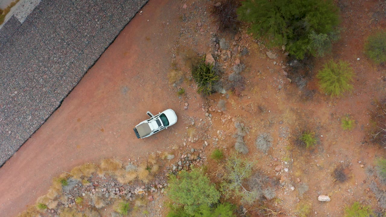 Overhead view in Puerto Escondido, Baja California Sur, with the white car lower on the dirt path, the paved road forming a dark diagonal edge, and no visible people.