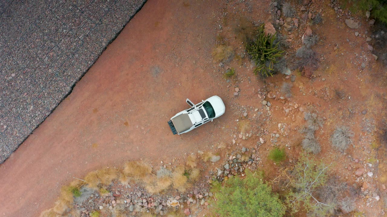Overhead frame from Puerto Escondido, Baja California Sur showing a white SUV near the upper road bend, red dirt, scattered rocks, sparse shrubs, a paved road edge, and no visible people.