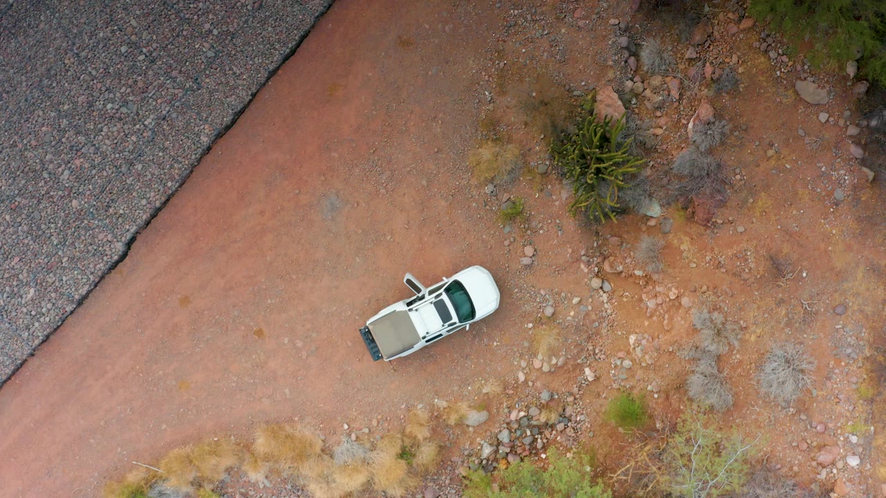 Aerial frame from Puerto Escondido, Baja California Sur showing the white SUV centered on red soil, with gravel, scrub vegetation, a paved road strip, and no visible people.