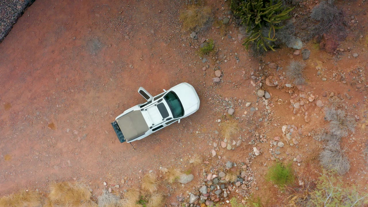 A white SUV sits near the left side of an overhead view of red rocky dirt in Puerto Escondido, Baja California Sur, with scattered stones, dry shrubs, green vegetation, and no visible people.