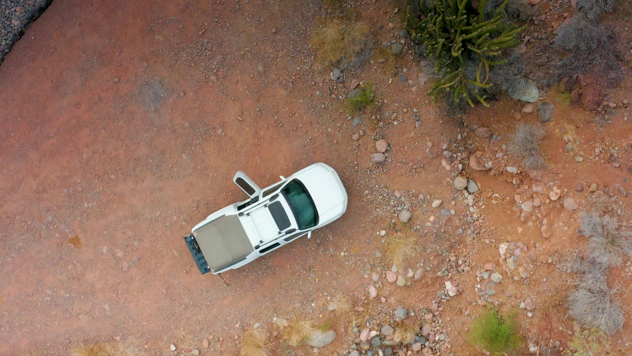 A top-down frame shows the white SUV progressing over red rocky ground in Puerto Escondido, Baja California Sur, with gravel patches, scattered stones, desert plants, and no visible people.