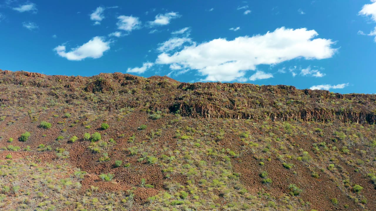 Second frame shows the reddish-brown slope in San Javier with sparse green shrubs, a dark ridge band, broad blue sky, shifted white clouds, and no visible people.