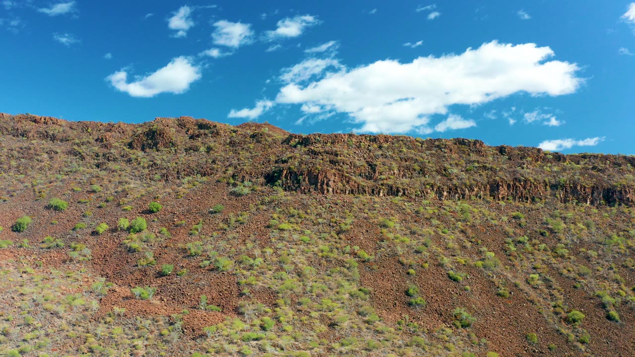 A reddish rocky hillside in San Javier, Baja California Sur sits below a bright blue sky with white clouds gathered near the upper center, with no visible people.