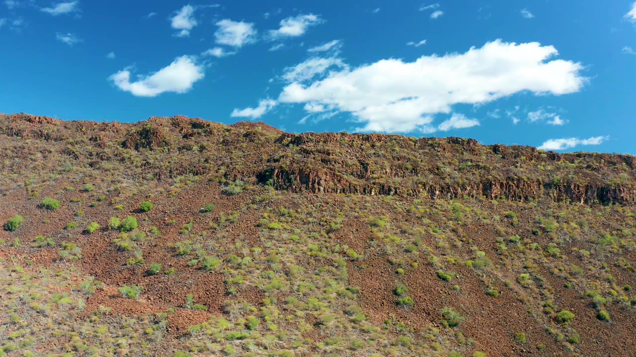 A jagged rocky ridge runs across the upper slope in San Javier, Baja California Sur, with small shrubs below and a broad white cloud over the blue sky, with no visible people.