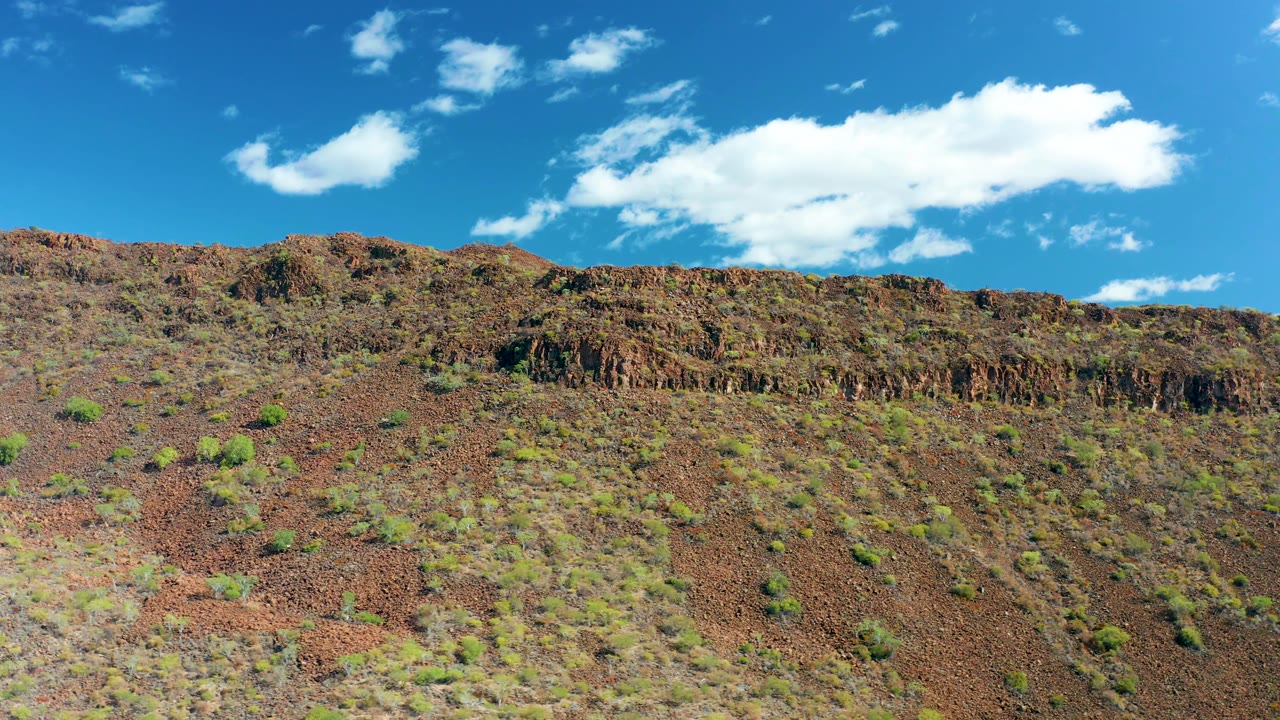 Red-brown hillside with green shrubs beneath scattered clouds.
