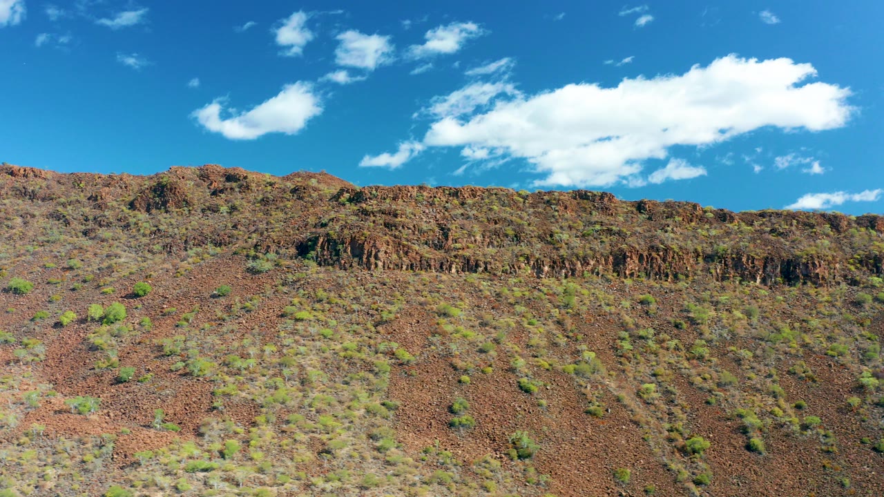 Wide view of a reddish-brown rocky hillside in San Javier, Baja California Sur, with scattered green shrubs, a ridge line, blue sky, white clouds, and no visible people.