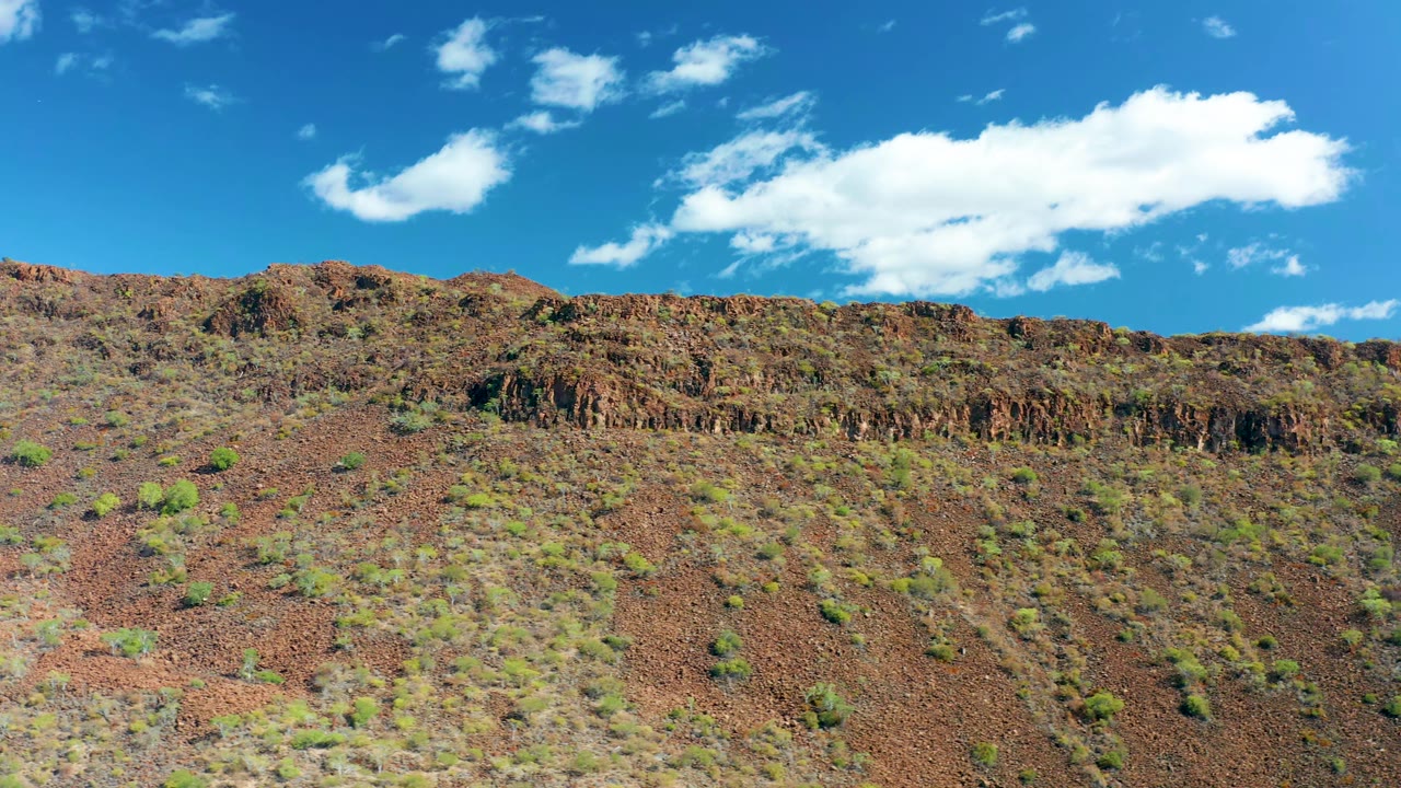 San Javier hillside frame with reddish rocky ground, scattered green vegetation, a dark craggy ridge across the middle, bright blue sky, a large white cloud, and no visible people.