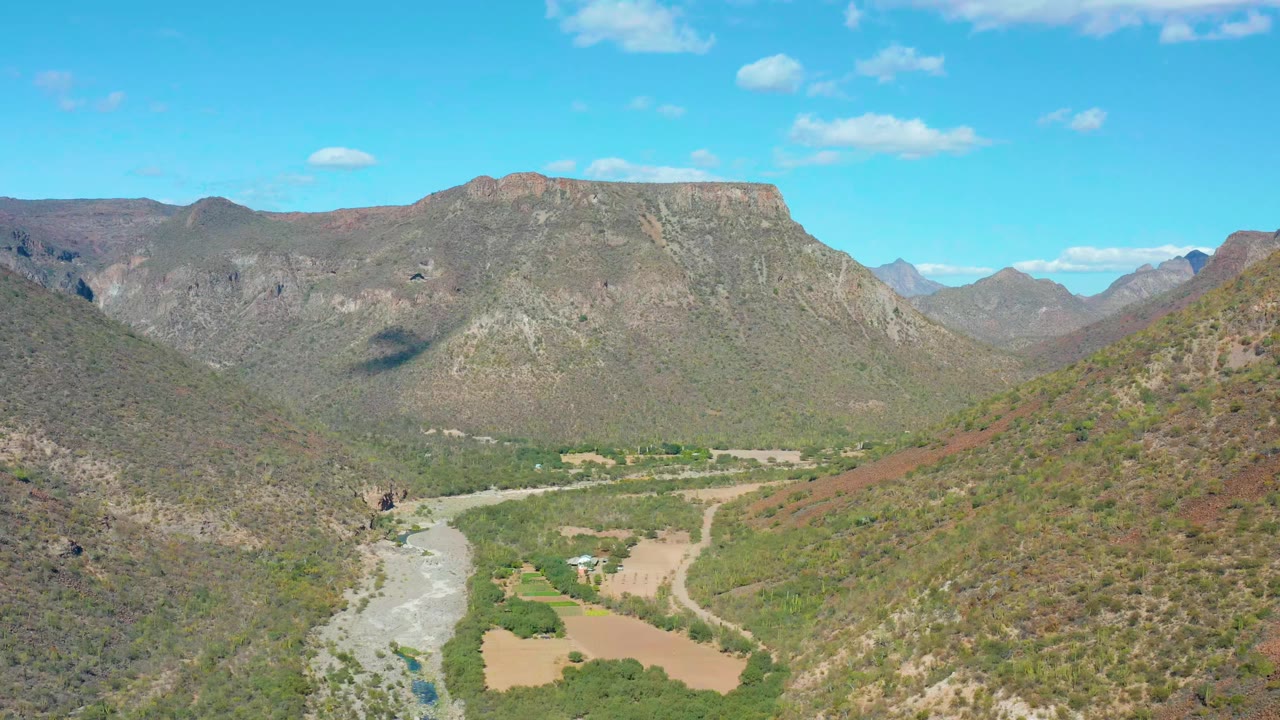 Early frame showing San Javier's river valley from above, with the river low in frame, scrubby hills on both sides, distant rocky peaks, scattered small clouds, and no visible people.