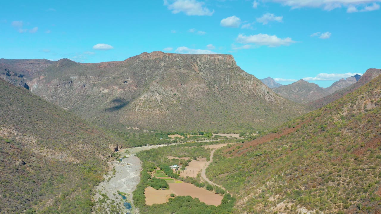 Mid-clip frame from above San Javier, Baja California Sur showing the river running along the lower left valley, green vegetation near the water, brown hillside slopes, high rocky mountains, and no visible people.
