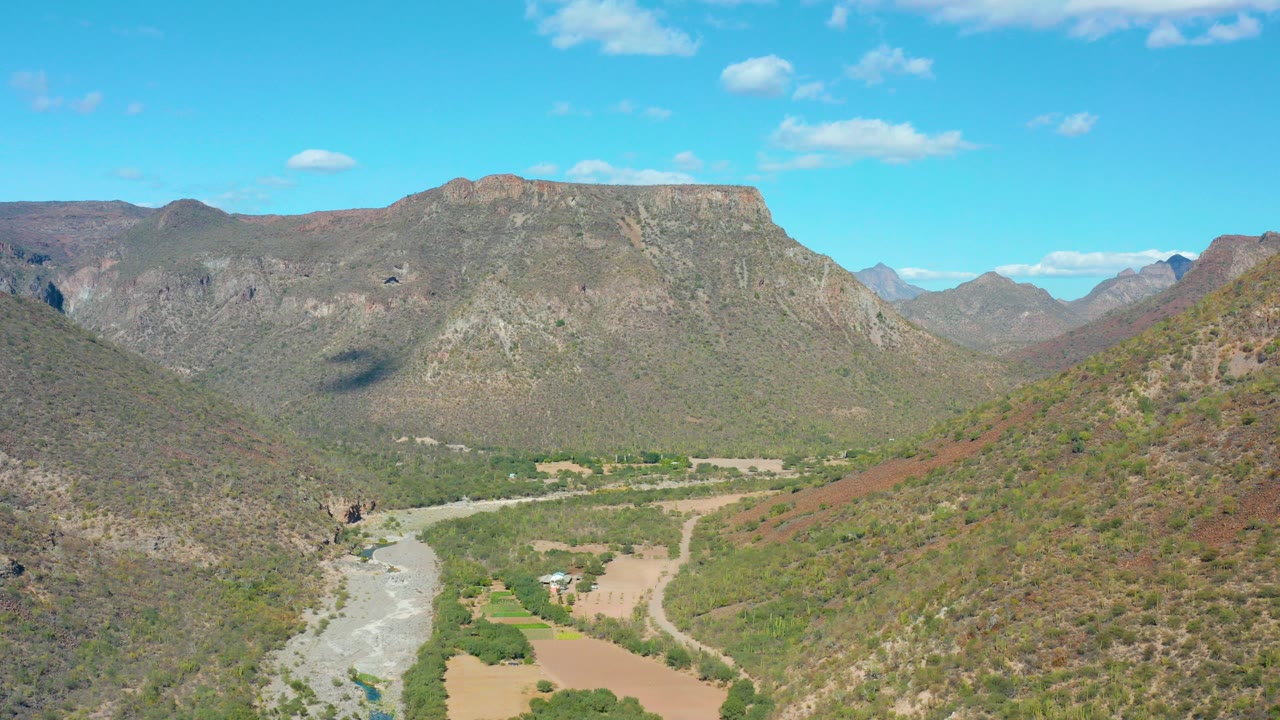 Opening aerial frame over San Javier, Baja California Sur, showing a pale river curving through a dry mountain valley under blue sky with no visible people.